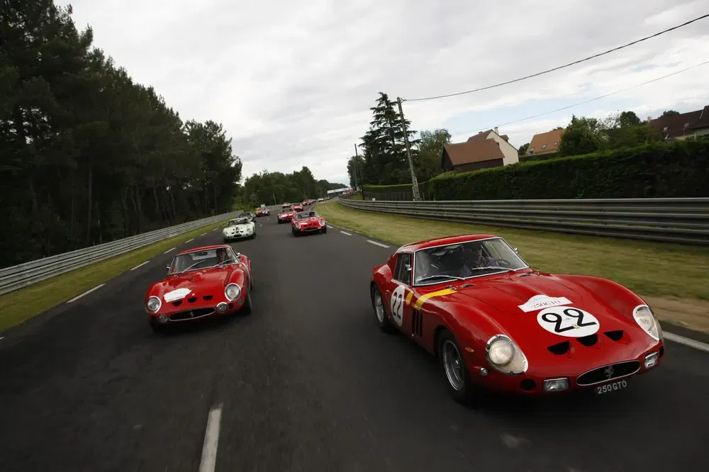 Ferrari 250 GTO rouges en course sur une route bordée d'arbres.