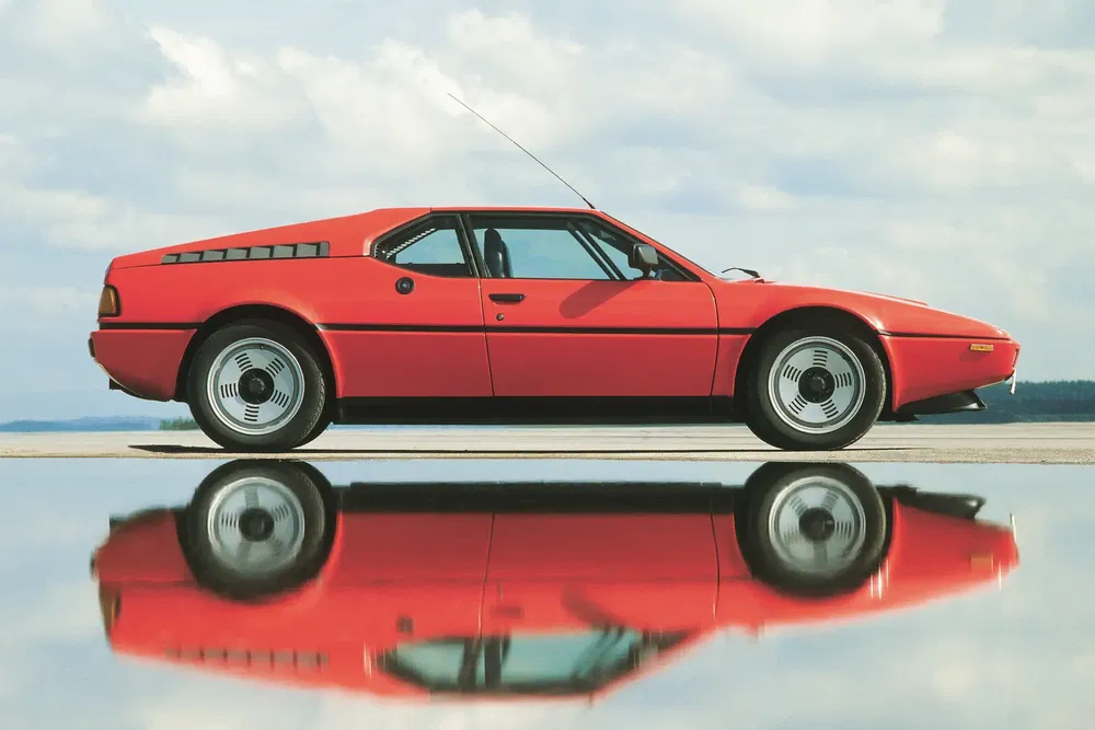 Voiture classique BMW M1 rouge avec reflet sur l'eau, design sportif et rétro.