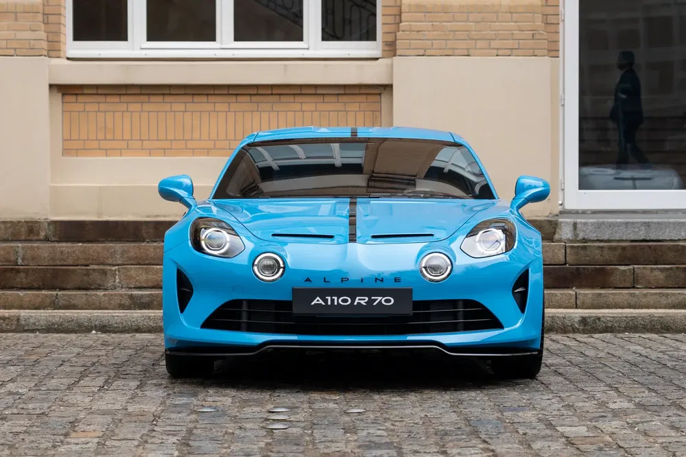 Vooraanzicht van blauwe Alpine A110 R70, geparkeerd voor een bakstenen gebouw.