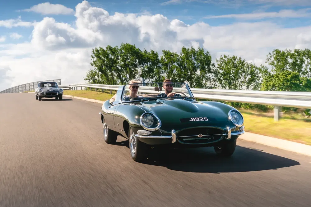 Twee groene Jaguar E-Types op zonnige weg met blauwe lucht op de achtergrond.