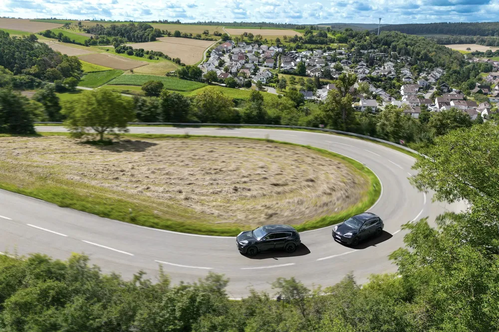 Deux voitures Tesla noires sur une route sinueuse, paysage verdoyant et village rural en arrière-plan.