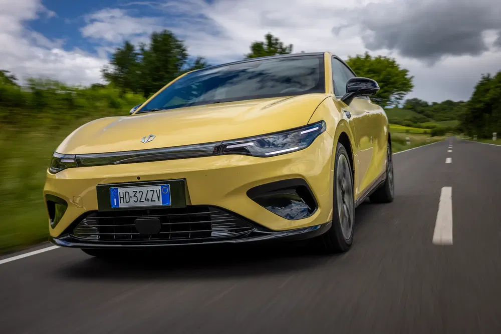 Voiture électrique jaune sur route, feux allumés, sous ciel nuageux et paysages verdoyants.
