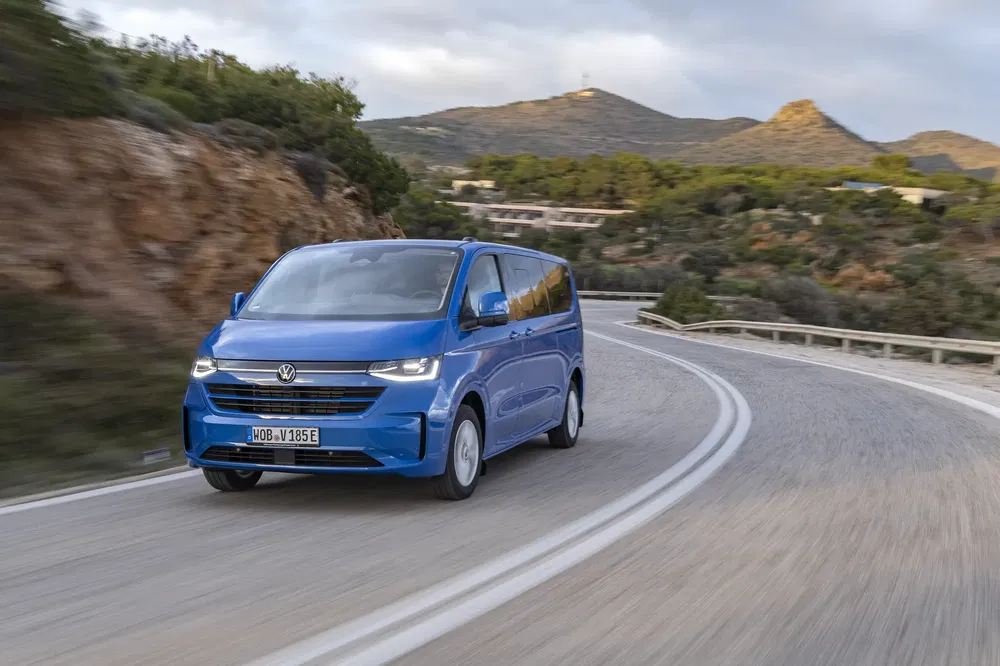 Blauwe VW Multivan op bochtige bergweg, biedt ruime transportmogelijkheden.