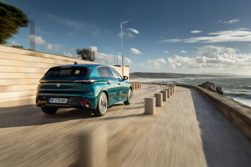 Blauwe Peugeot 308 rijdt op zonnige kustweg, omgeven door zee en barrières.