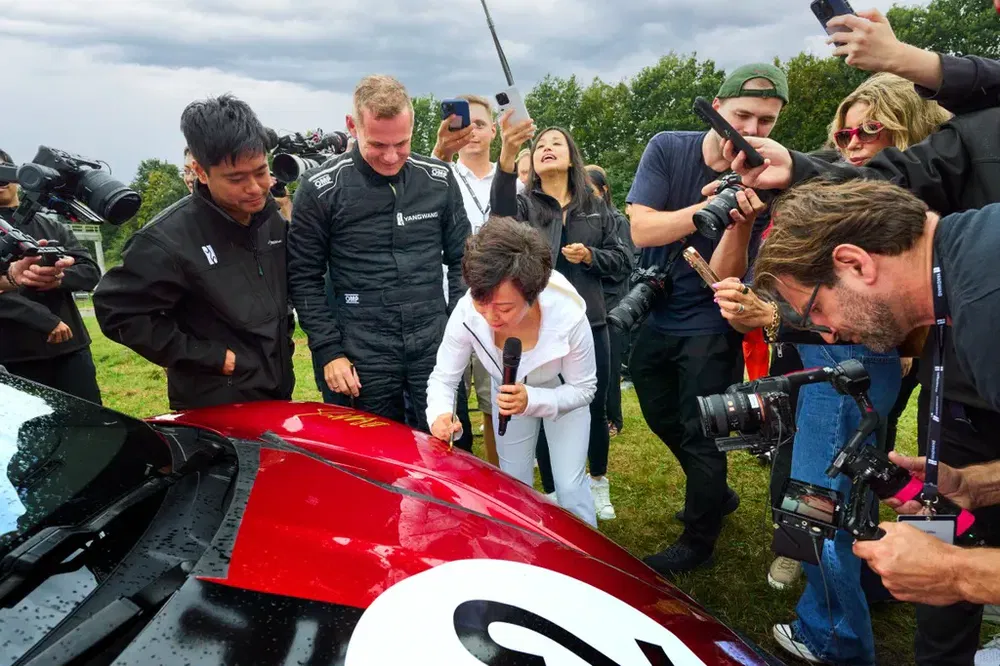 Persoon signeert rode raceauto, omringd door fotografen en journalisten.