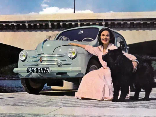 Une femme souriante assise près d'une Renault 4CV avec un chien noir à ses côtés.
