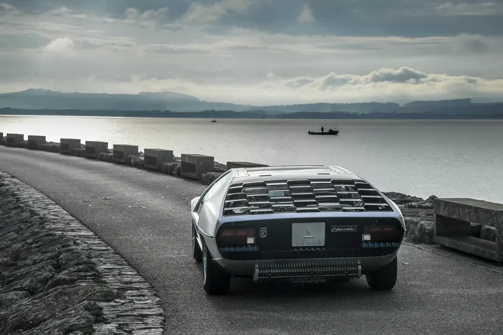 Zilveren Lamborghini Espada op een kustweg langs een meer onder bewolkte hemel.