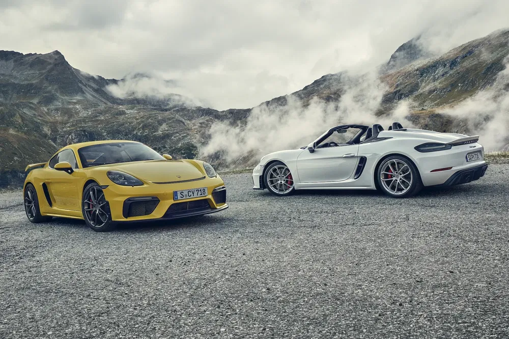 Deux Porsche 718, jaune et blanche, stationnées sur fond de montagne nuageuse.