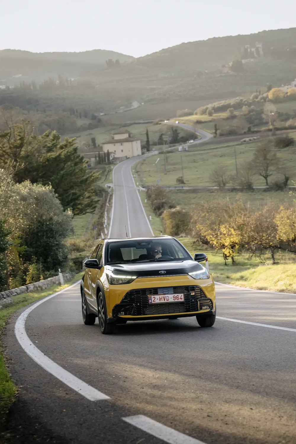 Toyota Aygo X jaune sur route sinueuse en campagne vallonnée, journée d'automne.