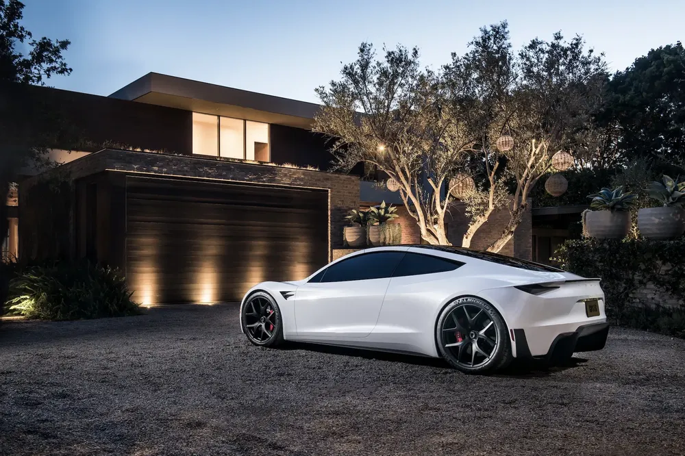 {"fr":"Tesla Roadster blanche garée devant une maison moderne illuminée la nuit.","nl":"Witte Tesla Roadster geparkeerd voor modern verlicht huis in de avond."}