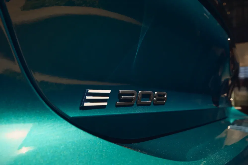 Gros plan sur l'emblème Peugeot E-308 sur une voiture électrique bleu métallisé.