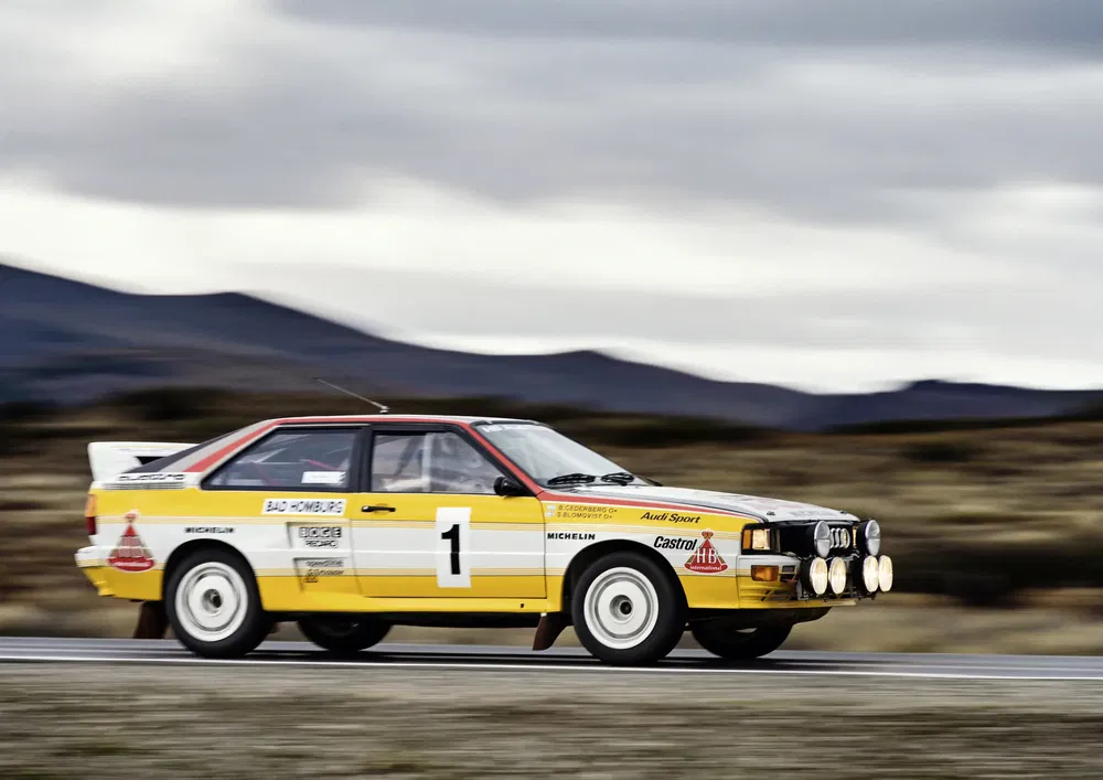 Audi Quattro jaune et blanche en course sur une piste avec ciel nuageux en arrière-plan.