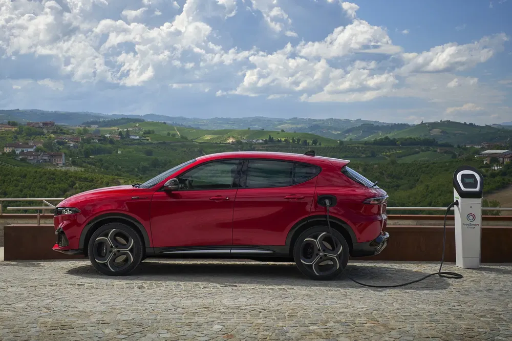 Rode Alfa Romeo Tonale SUV aangesloten op laadpaal met heuvelachtig landschap op de achtergrond.