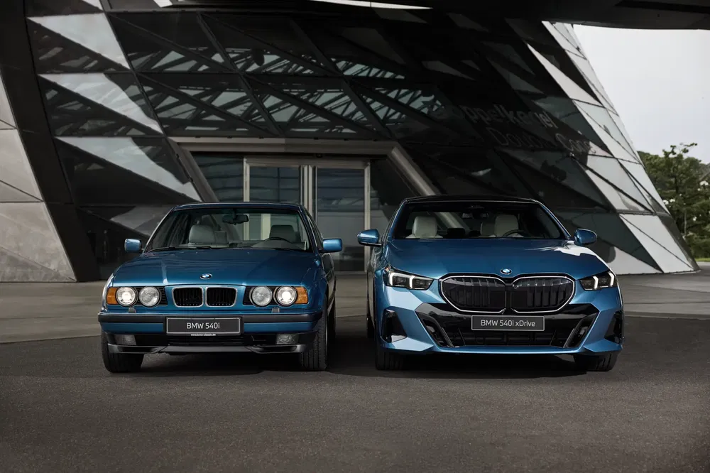 Twee blauwe BMW 540i auto's, oud en nieuw, voor een modern gebouw.