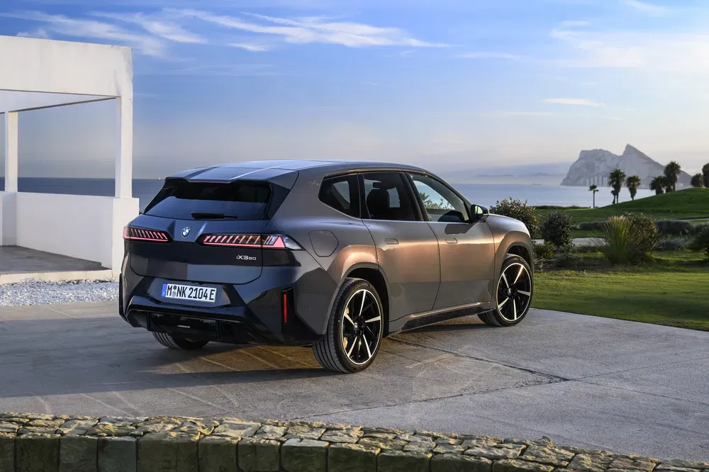 Grijze BMW iX3 aan kust met panoramisch uitzicht en blauwe lucht.