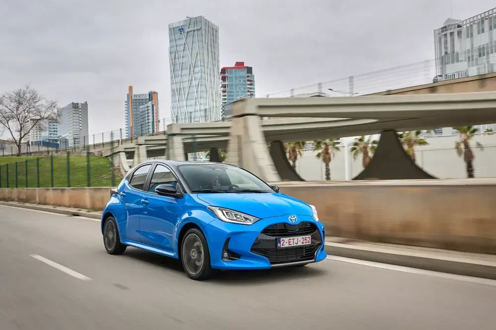 Toyota Yaris bleue roulant sur une route urbaine avec gratte-ciels en arrière-plan.