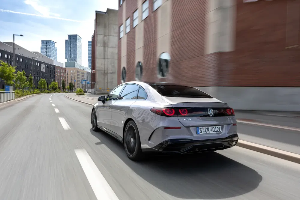 Zilveren Mercedes-Benz EQE rijdt in stedelijke omgeving, moderne gebouwen op de achtergrond.