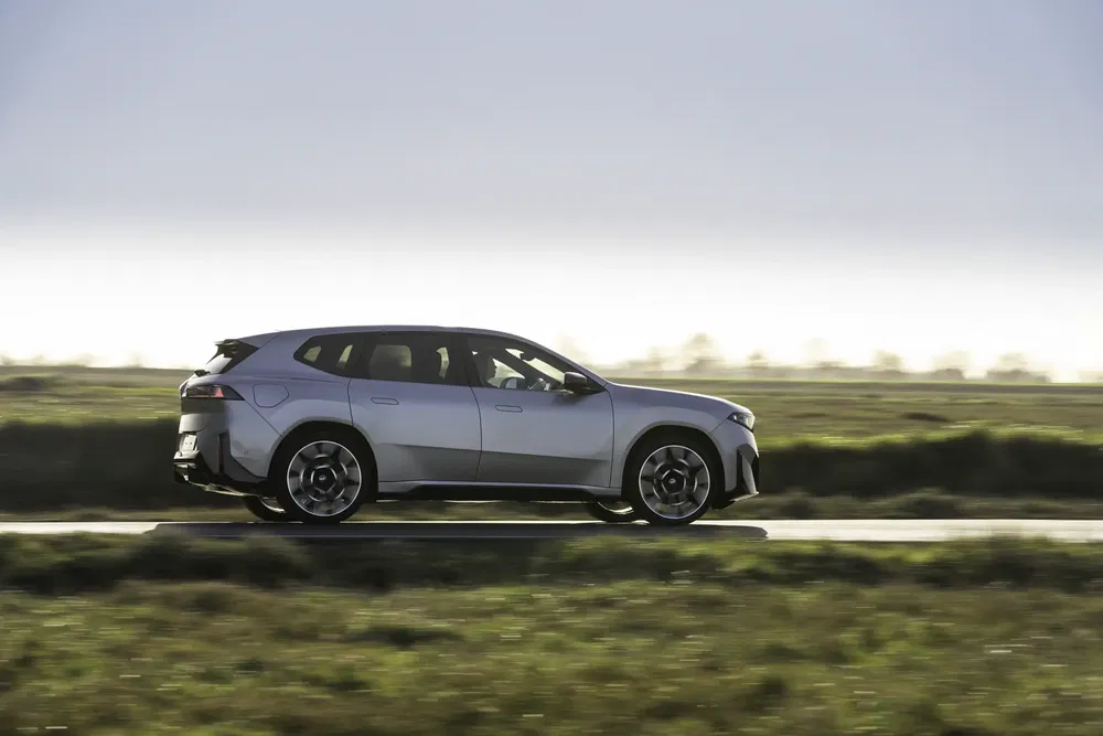 BMW iX en mouvement sur une route de campagne, flanc latéral sous un ciel clair.