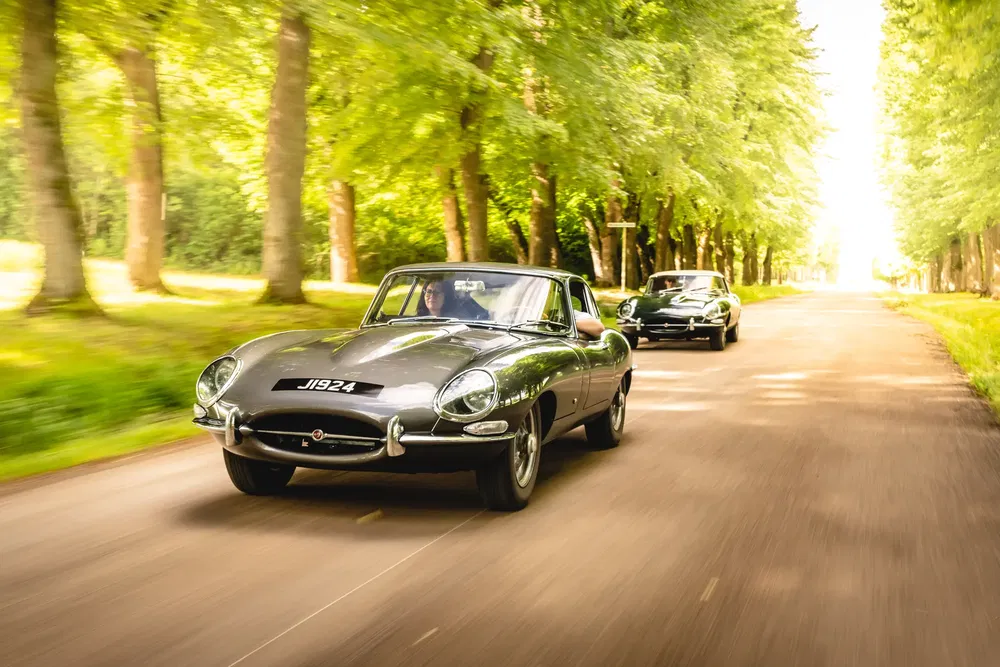 Klassieke Jaguar E-Type rijdt over boomrijke weg, elegante retro-sfeer.
