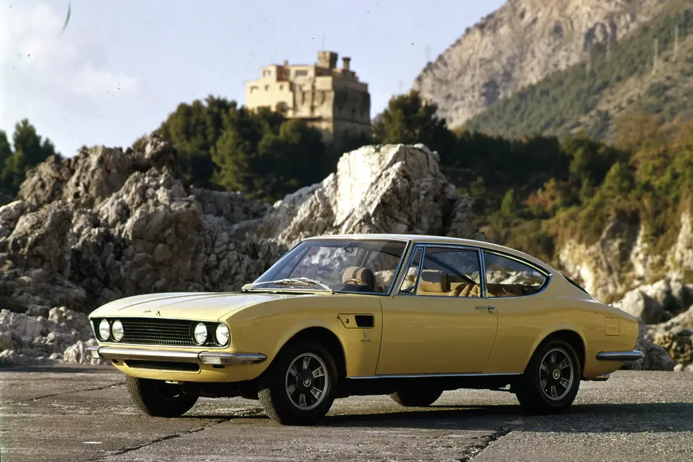 Fiat Dino Coupé jaune devant des montagnes rocheuses, style italien des années 1970.