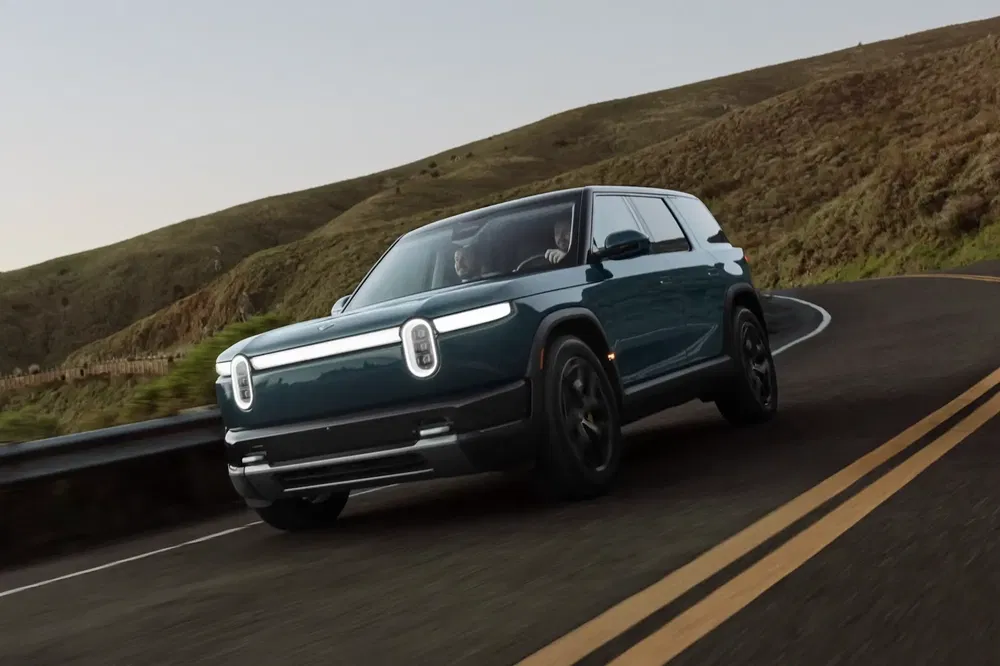 SUV électrique Rivian R1S roulant sur route sinueuse, entouré de collines verdoyantes.