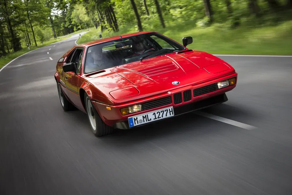 BMW M1 rouge classique roulant sur une route de campagne bordée d'arbres.
