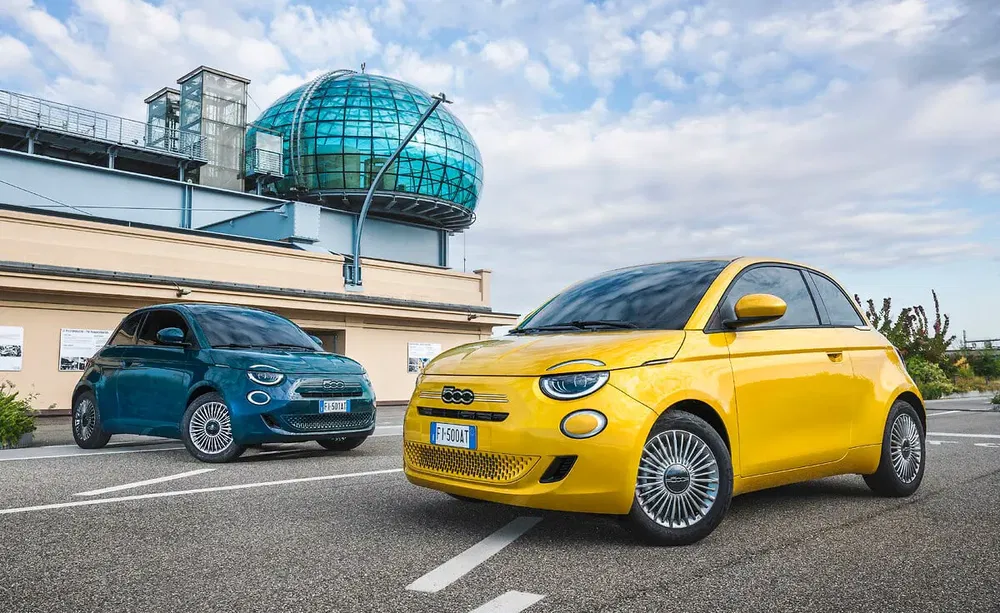 Twee elektrische Fiat 500, geel en blauw, geparkeerd voor een modern gebouw.